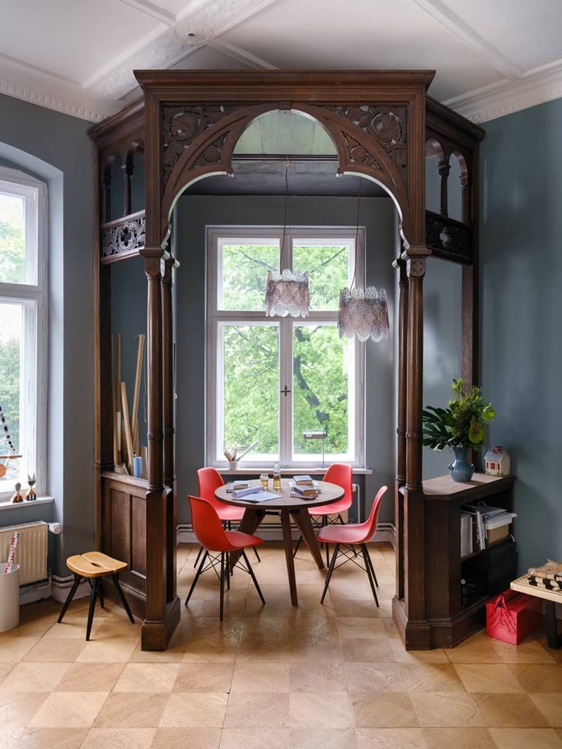 Holzerker mit geschnitztem Bogen und rundem Tisch mit roten Eames Plastic Chairs vor einem Fenster ins Gr&uuml;ne